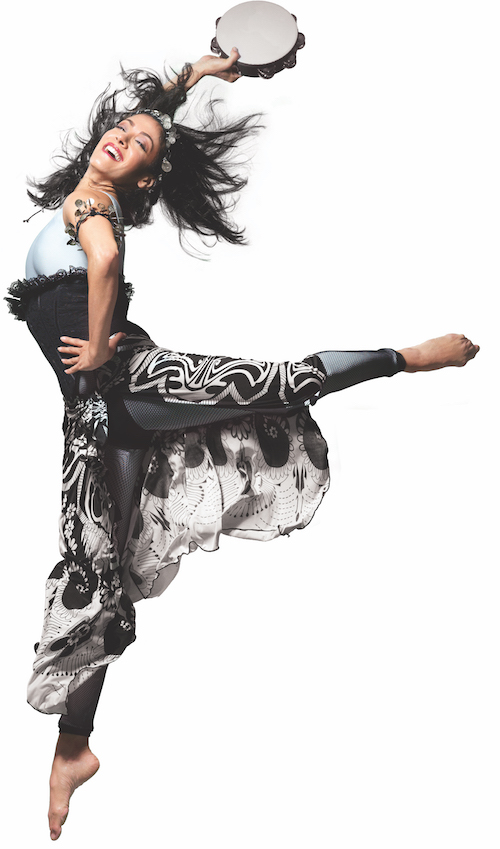 Parisa with her hand on her hip kicks her left leg behind her in an attitude position. She wears a gypsy inspired costume.